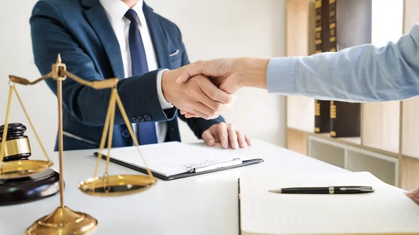 A personal injury attorney in a suit is shaking hands with a client across a desk beside the scales of justice and a gavel representing legal representation to help clients record trap evidence and build a strong case at Gusdorff Law P.C. in California.